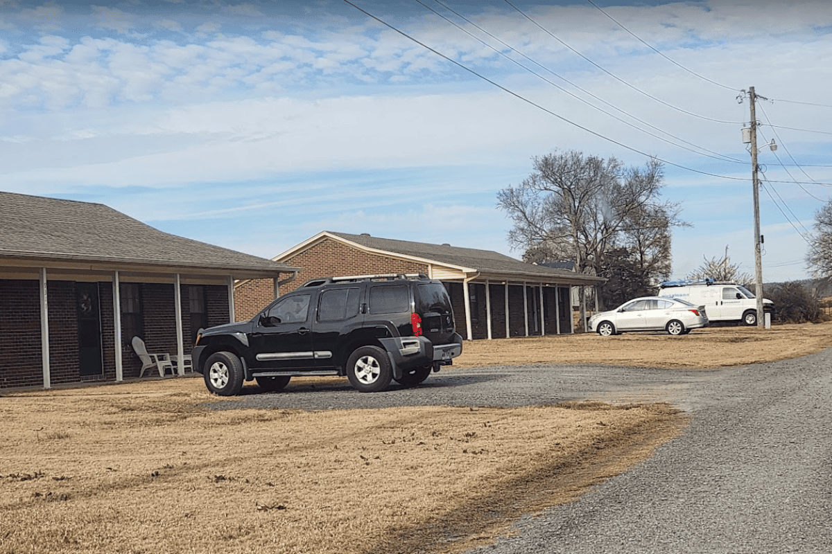A black SUV and two other vehicles are parked on a gravel lot in front of two brick buildings.